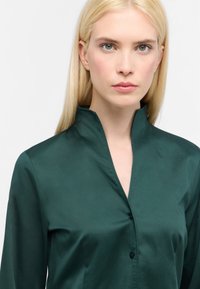 Grøn satinbluse med høj krave, som har knaplukning foran og lange ærmer. Glat tekstur med en subtil glans.