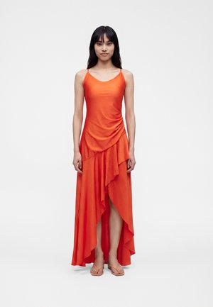 Femme debout portant une robe orange sans manches avec un ourlet volanté asymétrique et des sandales beiges sur un fond blanc uni.