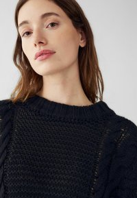 Sieviete ar garām brūnām matiem, tērpusies tumšā adītā džemperī, skatās nedaudz uz leju ar neitrālu izteiksmi uz fona, kas ir gaiši vienkrāsains.