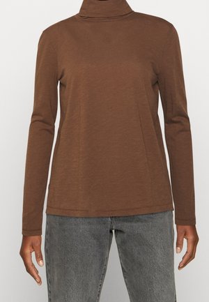 Chemise marron à col roulé manches longues portée avec un jean gris, montrant le haut du corps et les bras d'une personne sur un fond uni.
