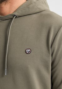Sudadera verde oliva con capucha ajustable, puños de canalé y un pequeño parche de logo circular en el pecho con colores contrastantes.