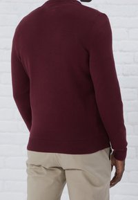 Pull en maille bordeaux avec un col rond, des poignets et un ourlet côtelés. Porté avec un pantalon beige clair. Le textile semble lisse et ajusté.