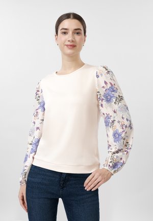 Νεαρή γυναίκα με σκούρα μαλλιά φοράει μπλούζα εκρού με φουσκωτά μανίκια με φλοράλ σχέδιο και μπλε τζιν, στέκεται μπροστά σε απλό φόντο.