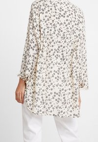 Kevyt, pitkähihainen beige pusero mustalla kukkakirjailulla, jossa pyöreä kaula-aukko ja rypytetyt hihat.