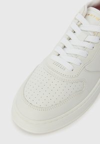 Baltas ādas sneakers ar perforētu pirkstu daļu, plakaniem baltajiem šņorēm un smalku zelta logotipu uz mēles. Gluda tekstūra un noapaļota forma.