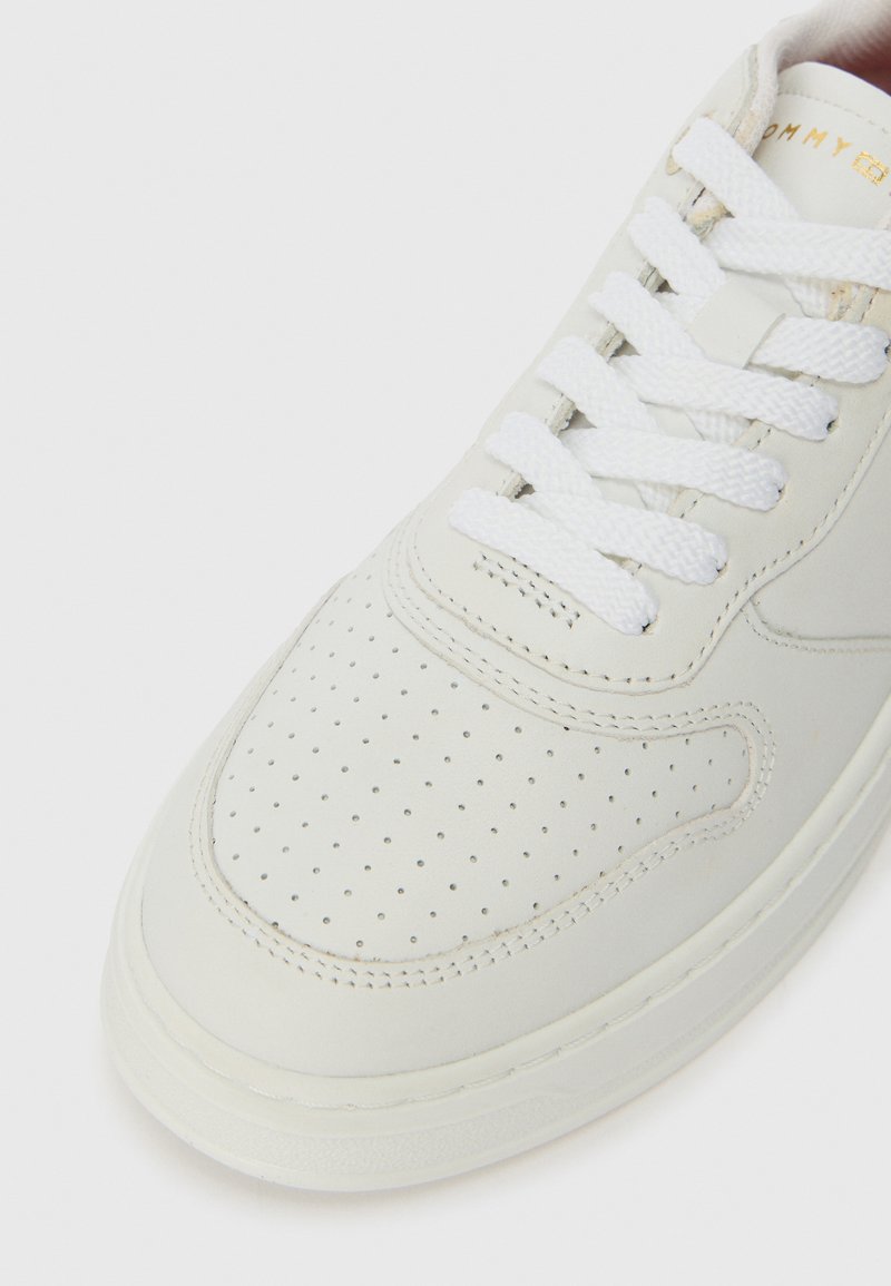 Zapatilla de cuero blanco con puntera perforada, cordones blancos planos y un sutil detalle de logo en dorado en la lengüeta. Textura suave y forma redondeada.