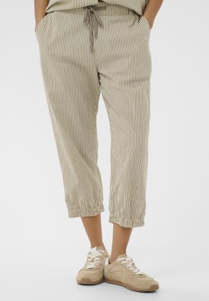 Osoba nosiaca béžové a biele vertikálne pruhované krátke nohavice s elastickými manžetami a béžové tenisky s bielymi šnúrkami. Ruky v vreckách.