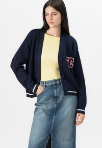 Σκούρο μπλε πλεκτό cardigan με κόκκινο κεντημένο λογότυπο 'SC', λευκές ρίγες στους καρπούς και χαλαρή εφαρμογή, σε συνδυασμό με κίτρινο τοπ και τζιν φούστα.