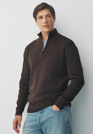 Brauner Baumwollpullover mit hohem Kragen und halbem Reißverschluss. Hat gerippte Bündchen und Saum; getragen über einem hellblauen T-Shirt und Jeans.