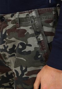 Kamelia-housut vihreän, ruskean ja mustan sävyissä, joissa on nappikiinnitys ja sivutaskut, valmistettu kestävästä kankaasta.