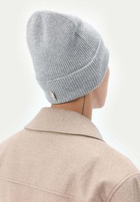 Ljusgrå ribbad stickad mössa med avsmalnande topp. Har en liten etikett på sidan. Bärs med en beige yllekappa.