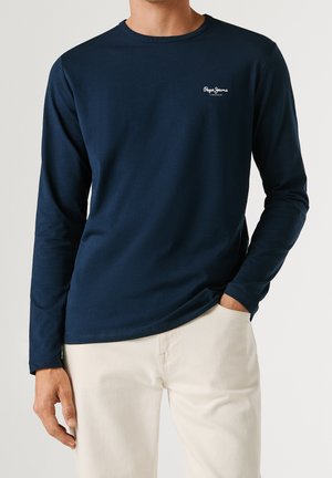 Homme portant une chemise à manches longues bleu marine avec un petit logo blanc "Pepe Jeans London" sur la poitrine et un pantalon beige clair, main dans la poche.
