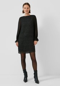 Czarna sukienka z teksturowanej tkaniny i przezroczystymi długimi rękawami, o prostym kroju i falbaniaste mankiety, zestawiona z czarnymi botkami do kostki.