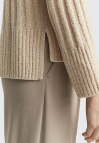 Beige geribbelde gebreide trui met een splitzoom, gecombineerd met licht taupe slim-fit broek. De stof lijkt zacht en textuur te hebben.