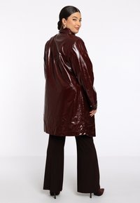 Femme dans un long manteau marron foncé brillant, portant un pantalon large noir et des chaussures à talons, se tient de dos, regardant par-dessus son épaule avec un léger sourire.
