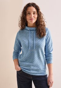 Ljusblå hoodie med ett strukturerat mönster, med en krage och dragsnören, långa ärmar och en avslappnad passform, ihop med mörka jeans.