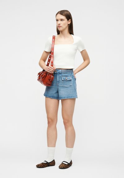 Γυναίκα που στέκεται με κόκκινη τσάντα ώμου, φορώντας λευκή μπλούζα με κοντά μανίκια, τζιν σορτς, λευκές κάλτσες και παπούτσια με λεοπάρ μοτίβο.