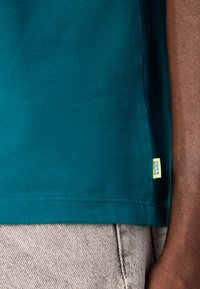 Søgrøn bomulds t-shirt med en kantmærke med logo, parret med lysegrå teksturerede stofbukser. Fokus på trøjens glatte tekstur og farve.