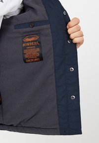 Binnenkant van een marine bomberjack met textuurstof, grijze paneel, drukknopen en een gelabeld label met merk en specificaties.