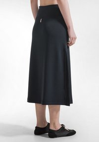 Mujer con falda midi negra con abertura lateral y zapatos planos negros con cordones, vista desde atrás sobre un fondo blanco liso.