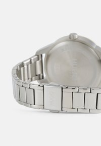 Reloj de acero inoxidable con una pulsera plateada elegante, que presenta una caja redonda simple y un cierre grabado con "HUGO". Acabado suave y pulido.