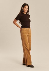 T-shirt marron ajusté à manches courtes associé à un pantalon en velours côtelé taille haute et à jambes larges, de couleur beige chaud. Chaussures noires.