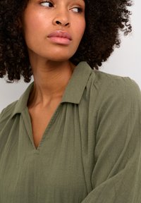 Blusa verde oliva con colletto, caratterizzata da un scollo a V e un tessuto morbido e texturizzato. Il dettaglio plissettato sulle spalle aggiunge profondità.