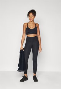 Sujetador deportivo negro y leggings de cintura alta hechos de tela elástica; el modelo sostiene una toalla oscura. Zapatillas deportivas negras completan el look.