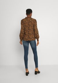 Blusa con stampa leopardata in marrone e nero, con colletto alto e maniche a sbuffo; abbinata a jeans slim blu scuro e sandali neri con tacco blocco.
