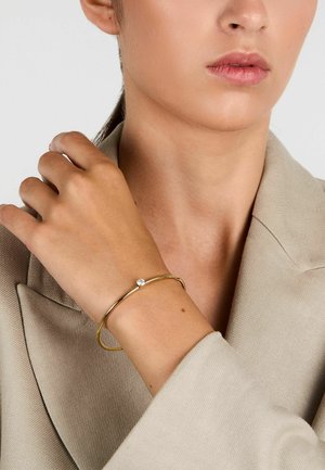 Bracciale rigido color oro con una finitura liscia e una pietra preziosa trasparente incassata, indossato su una mano con un blazer in tonalità neutra.