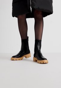 Svarte skinn ankelstøvler med en kraftig beige såle. Støvlene har en glatt overflate og en tettsittende sokkelignende åpning.