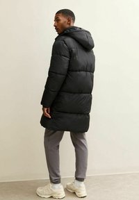 Hombre de pie con un abrigo acolchado negro con capucha, pantalones jogger grises y zapatillas beige, mirando por encima del hombro contra un fondo liso.