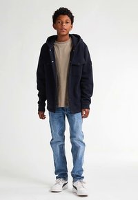 Jachetă bleumarin cu glugă, tricou bej, blugi albastru deschis, adidași albi. Jacheta are închidere cu nasturi și două buzunare frontale.