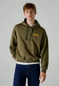 Sweatshirt verde oliva feito de material felpudo, com um bolso frontal e o logótipo amarelo "RBR". Combinado com jeans de denim azuis.