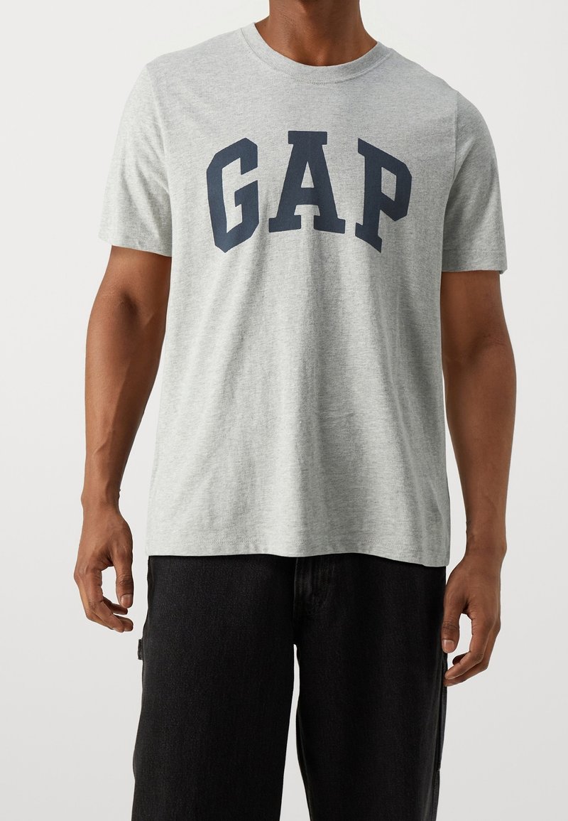 T-shirt en coton gris avec "GAP" imprimé en lettres capitales bleu foncé sur la poitrine. Il présente un col rond et des manches courtes.