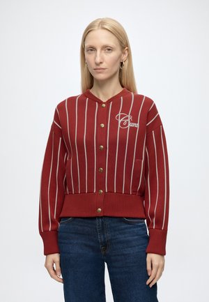 Vrouw met lang blond haar draagt een rood-wit gestreept, geknoopt vest met ""Candy"" op de borst, gecombineerd met blauwe jeans, staat neutraal.