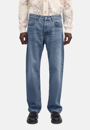 Uomo che indossa jeans larghi color azzurro chiaro, mocassini neri e un maglione crema con motivo astratto rosa, in piedi davanti a uno sfondo bianco.