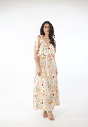 Mujer de pie que lleva un vestido largo sin mangas con estampado floral, tirantes anudados en los hombros y tacones abiertos beige sobre un fondo blanco.