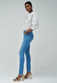 Blusa blanca con detalles de encaje, jeans ajustados azules y sandalias de tacón beige. La modelo está de lado frente a un fondo neutral.