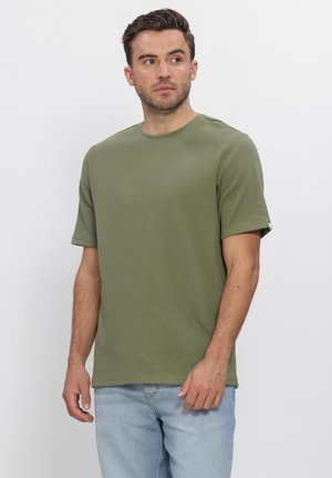 Man met kort bruin haar die een olijfgroen shirt met korte mouwen en lichtblauwe spijkerbroek draagt, kijkt naar links.