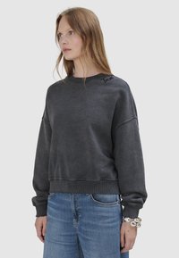 Frau mit langen Haaren trägt ein ausgewaschenes, schwarzes, oversized Sweatshirt mit eingesticktem Text auf der Schulter, blaue Jeans und ein massives silbernes Armband.