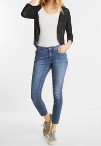 Svart lättviktskofta över en vit t-shirt, kombinerad med ljusblå smala jeans och grå sneakers med beige detaljer.