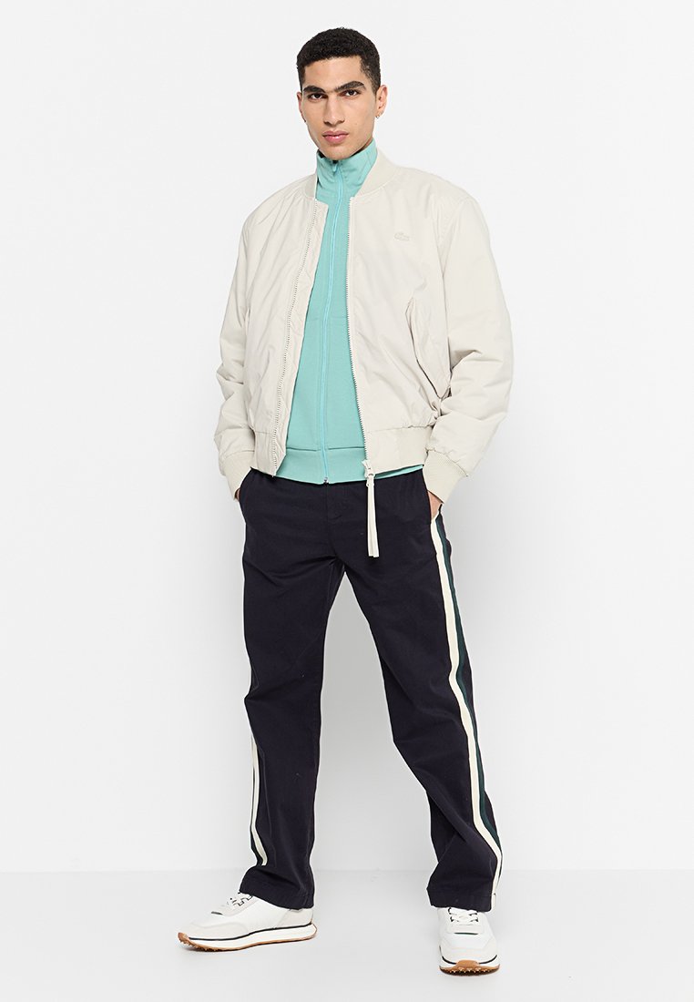 Bomber jacket beige chiaro sopra una maglia teal con zip, abbinata a pantaloni scuri con righe laterali. Il modello indossa scarpe da ginnastica bianche.