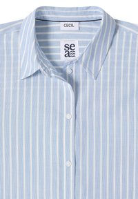 Chemise bleu clair à boutons avec rayures verticales blanches, col pointu et deux boutons visibles, avec les étiquettes "CECIL" et "sea" à l'intérieur du col.