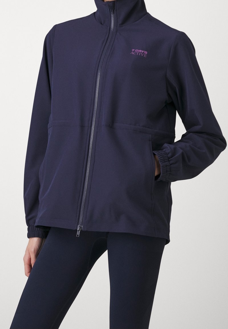 Personne portant une veste zippée bleu marine avec col montant, poignets élastiques, poches latérales et logo "7 Days Active" sur la poitrine.