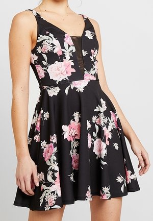 Femme portant une robe noire sans manches avec des motifs floraux roses et blancs et un décolleté en V profond avec une incrustation noire transparente.