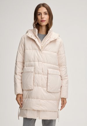 Kobieta ubrana w jasnobeżową puchową kurtkę do kolan z dwoma dużymi przednimi kieszeniami i wysokim kołnierzem, na szarych spodniach.