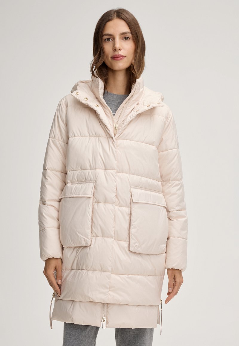 Femme portant un manteau matelassé beige clair mi-long avec deux grandes poches frontales et un col haut, sur un pantalon gris.