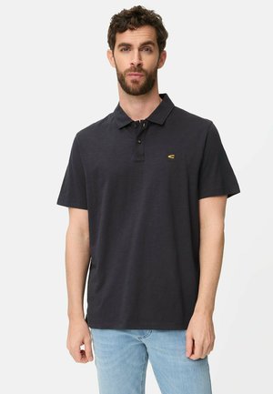 Mann mit kurzen lockigen Haaren, der ein schwarzes Polohemd und hellblaue Jeans trägt und vor einem schlichten weißen Hintergrund steht.