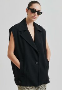 Svart ärmlös blazer i texturerad tyg, med stora slag, två framfickor och dubbelknappsstängning.